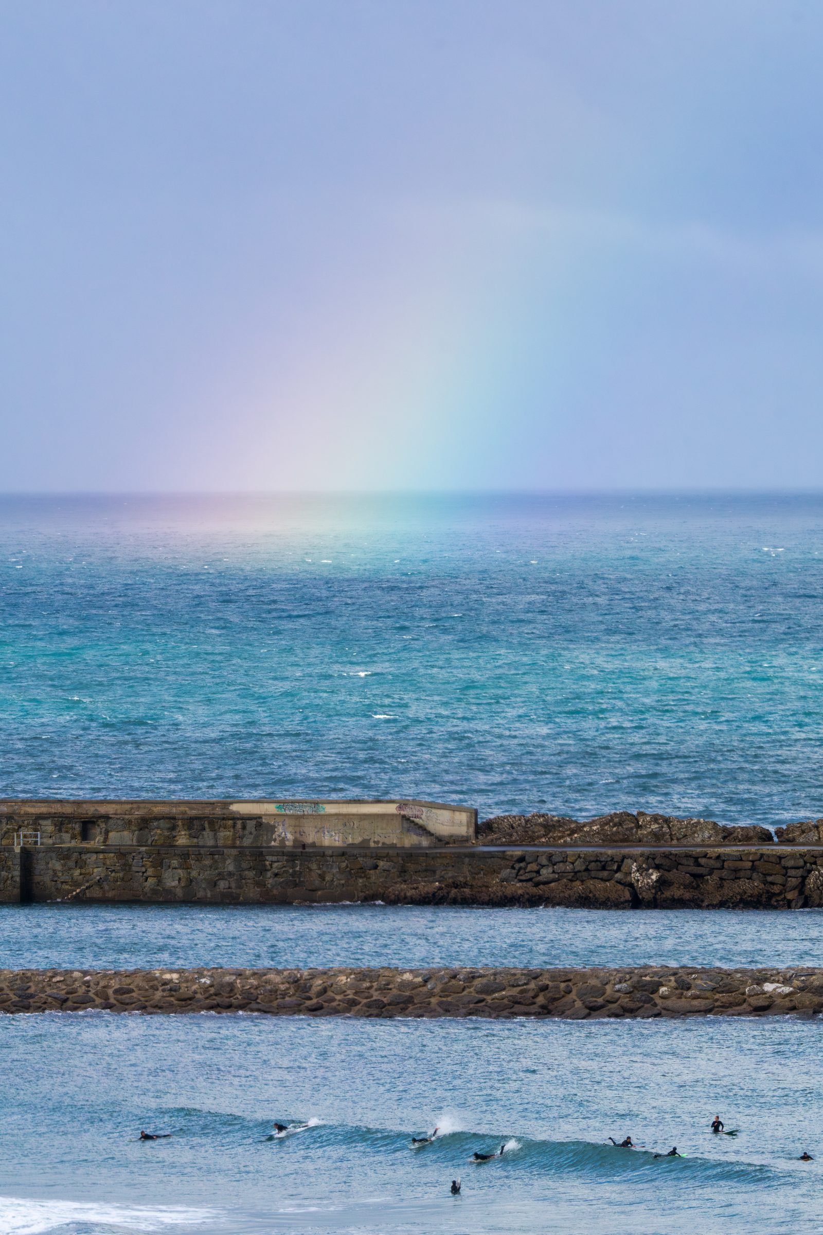 A faint rainbow melts into the sea as surfers drift together, patient and unhurried, finding quiet companionship in the rhythm of the waves.