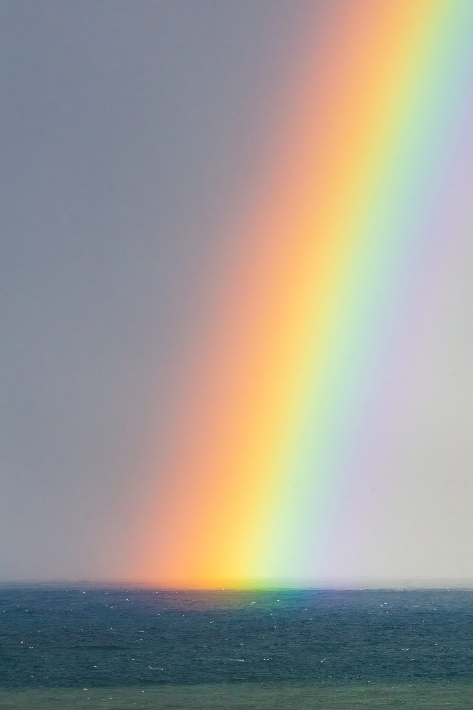 Where the sea meets the sky, a rainbow bends low to remind us that even in gray moments, color finds its way to touch the horizon.